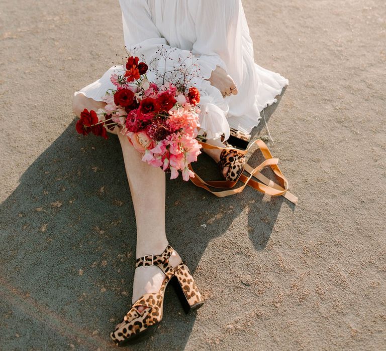 Bride in a short weddng dress with pearl encrusted birdcage veil, red lipstick and leopard print platform shoes holding a red wedding bouquet 