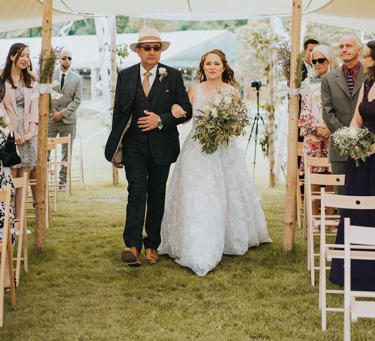 Bride in lace wedding dress holding white bouquet is walked down the aisle by father in blue checkered suit and white hate with yellow stripe 