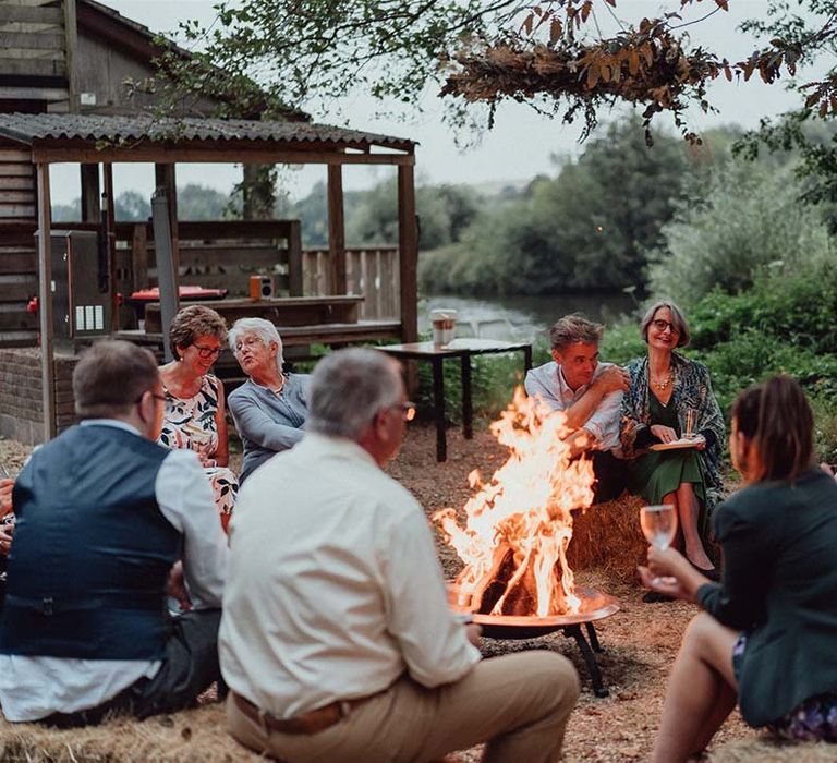 Guests sit around fire pit next to cabin near lake at wedding venue