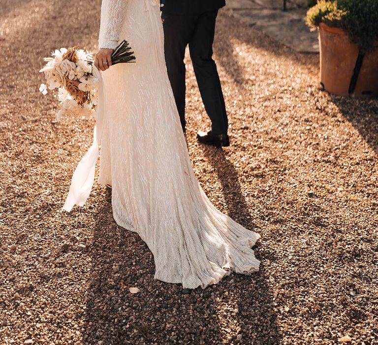 Bride looks at the camera in beaded dress holding neutral nude, toffee and brown wedding bouquet holding hands with groom with dried flower buttonhole