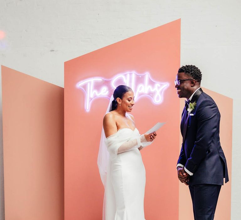 Smiling bride in off the shoulder fitted wedding dress reads out her vows to the groom in black tie