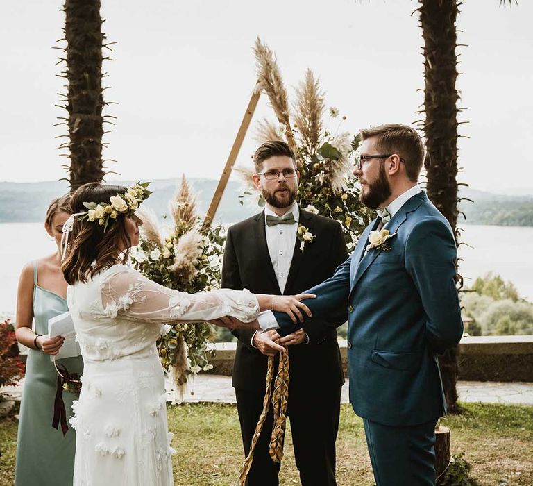 Outdoor wedding ceremony with bride in an appliqué wedding dress holding arms with her husband during the handfasting ceremony