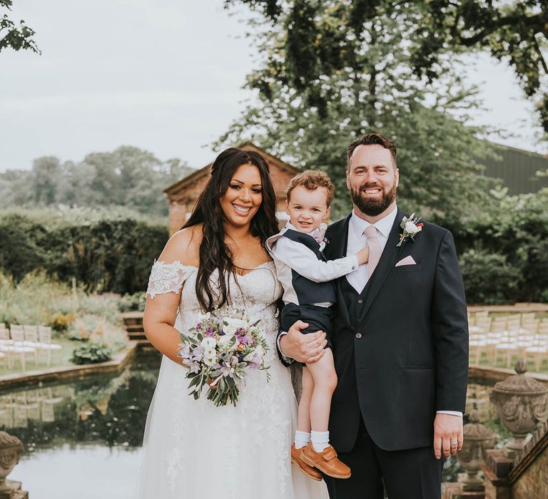 Bride in off the shoulder wedding dress with purple wedding bouquet stands with little page boy and groom in dark grey suits and pink ties 