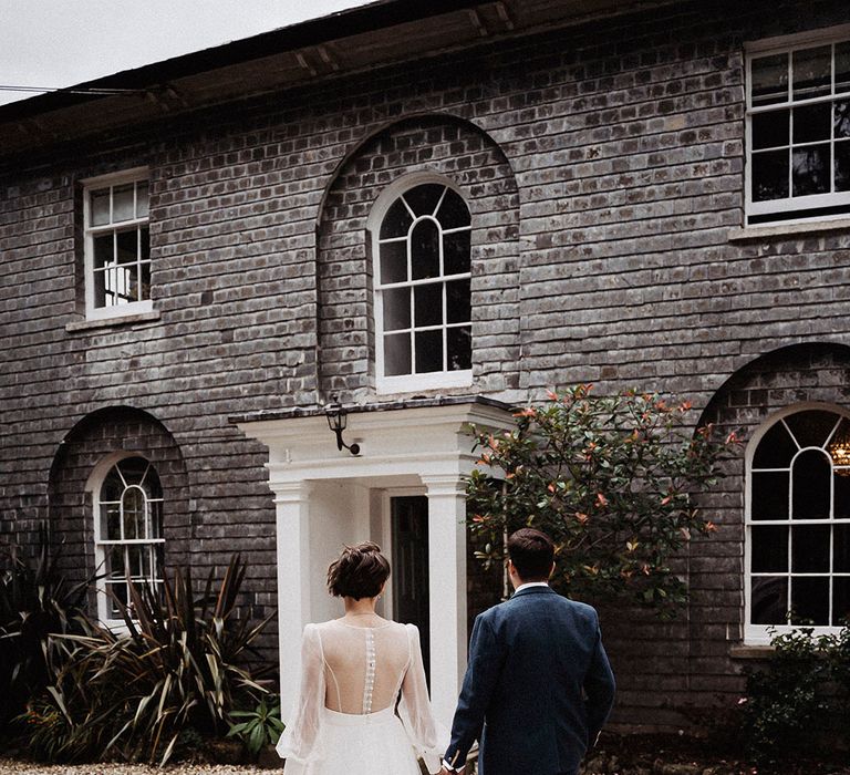 Bride in sheer long sleeve wedding dress holding hands with groom in blue suit walk toward their wedding venue, Treseren