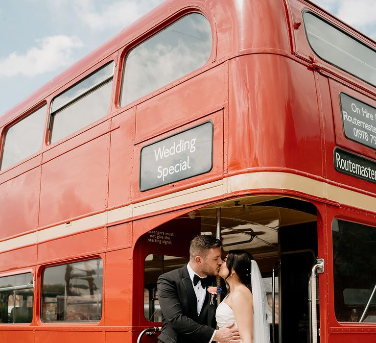 Bride in pearl veil and one shoulder dress kisses the groom in black tie standing on the bus wedding transport 