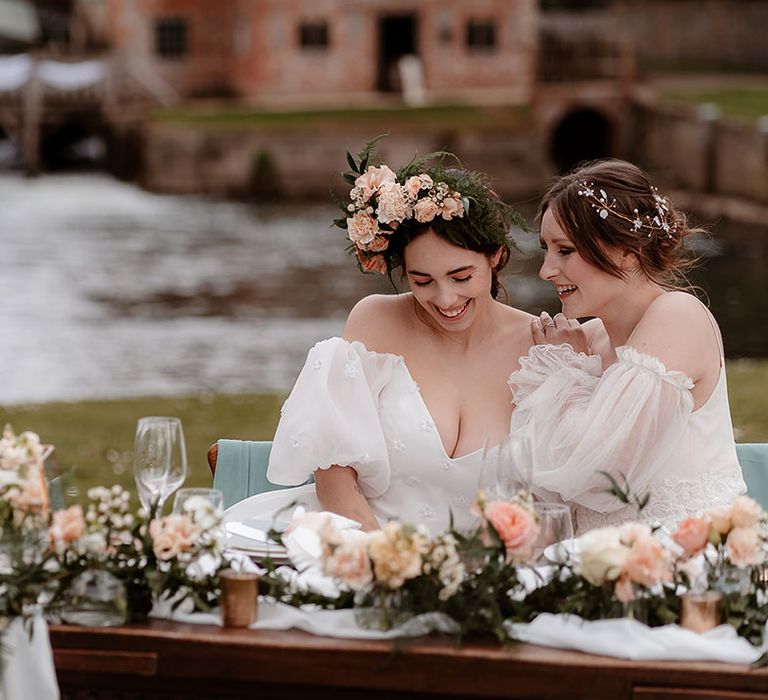 Two brides sitting at their outdoor sweetheart table decorated with drapes, pink flowers and lanterns in a floaty wedding dress with spaghetti straps and strapless wedding dress with puff sleeves