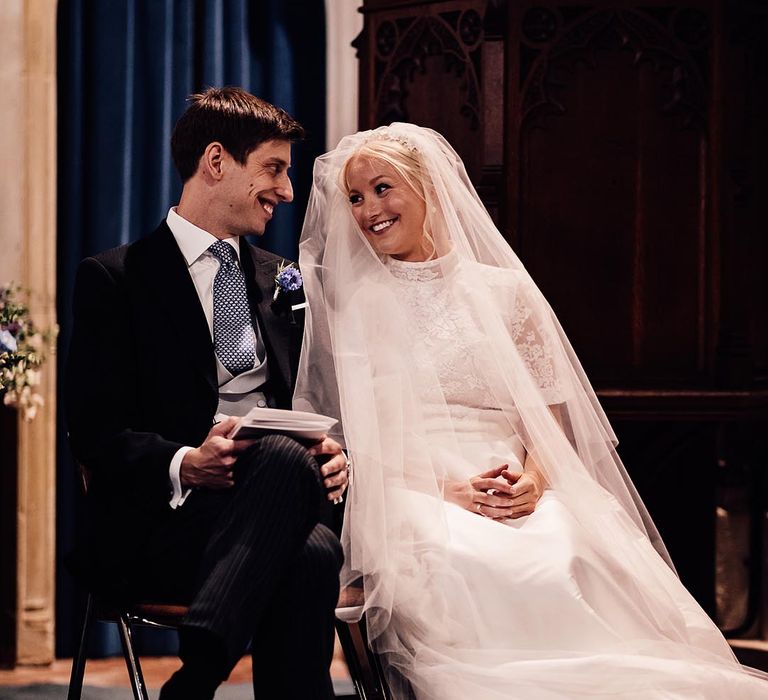 Groom in black suit with blue flower buttonhole and grey waistcoat sits smiling with bride in lace wedding dress during their ceremony 