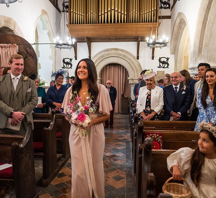 Bridesmaid walks down the aisle wearing pastel pink bridesmaid dress with 1/4 length sleeves and tie to front of waist