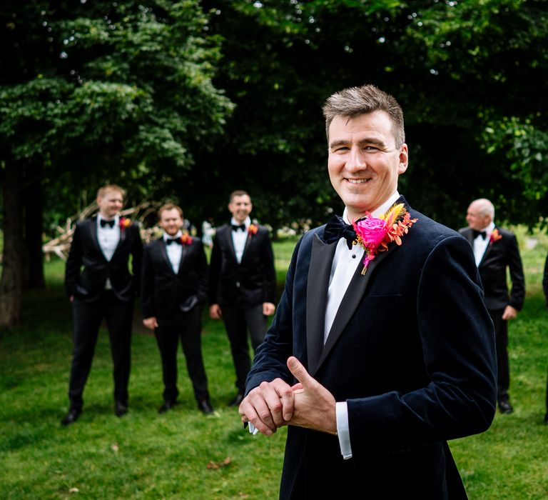 Groom wears blue velvet dinner jacket complete with black tie and colourful floral buttonhole as his groomsmen stand behind him