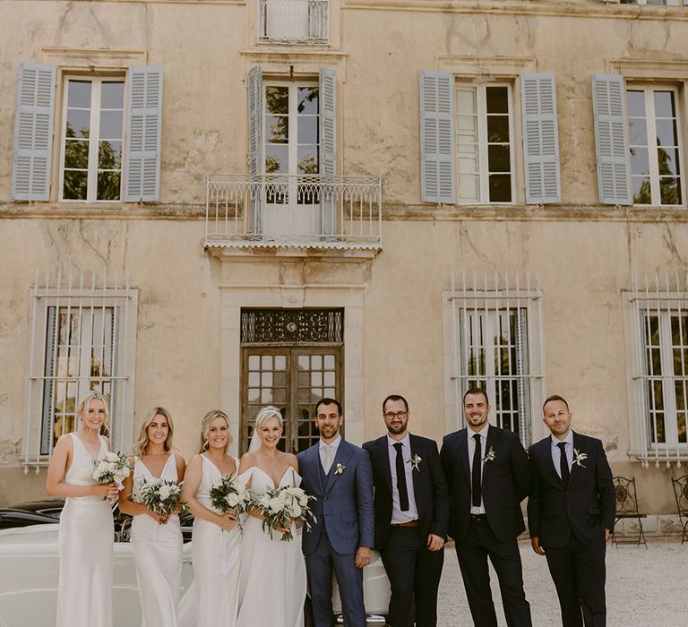 Bride & groom stand with their bridesmaids who wear white silk dresses and groomsmen in black suits with floral buttonholes 