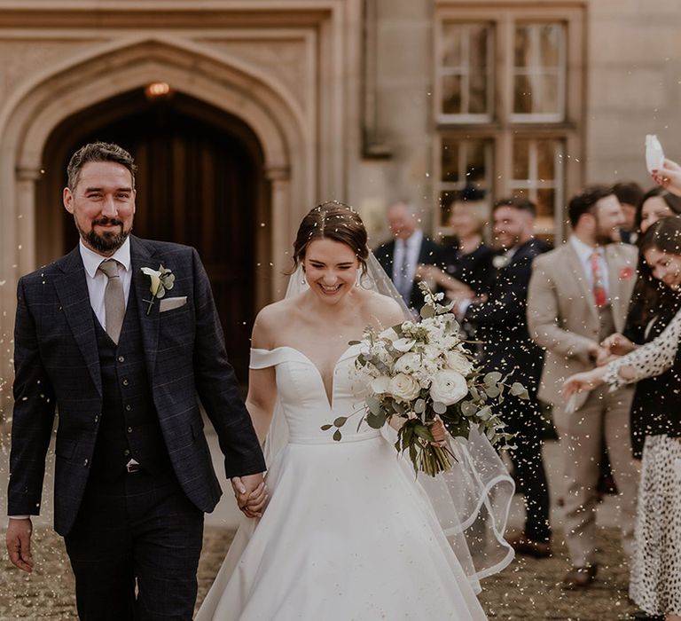 White confetti exit for the bride in an elegant off the shoulder wedding dress and groom in a deep blue suit 