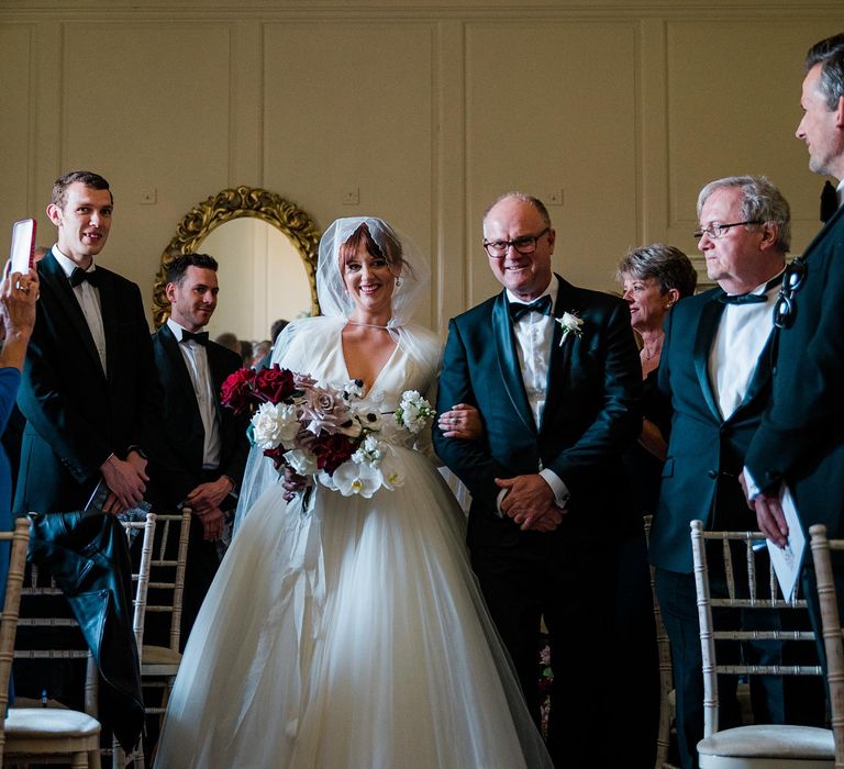 Bride walks down the aisle wearing princess tulle wedding dress with V-shaped neckline and cathedral veil whilst holding bright floral bouquet