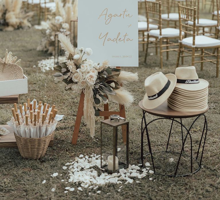 Umbrellas in wicker basket beside white hats and white welcome sign surrounded by pampas grass decor 