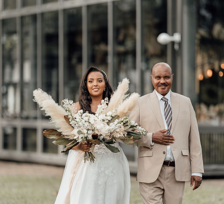 Black bride carries oversize pampas grass bouquet filled with green palm leaves and white roses as she walks down the aisle in Phuket 