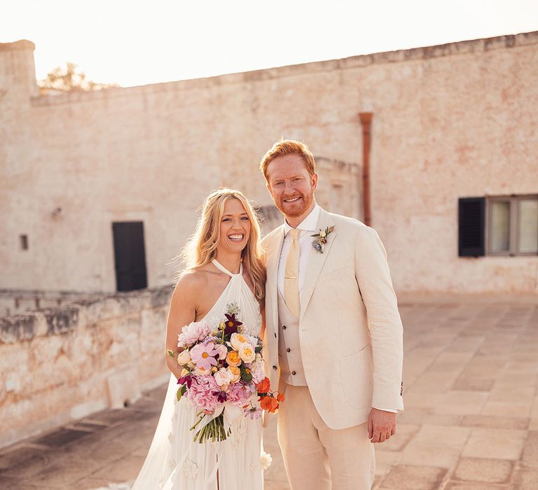 Bride holding a purple, red and pink wedding bouquet stands smiling with the groom in a cream suit 