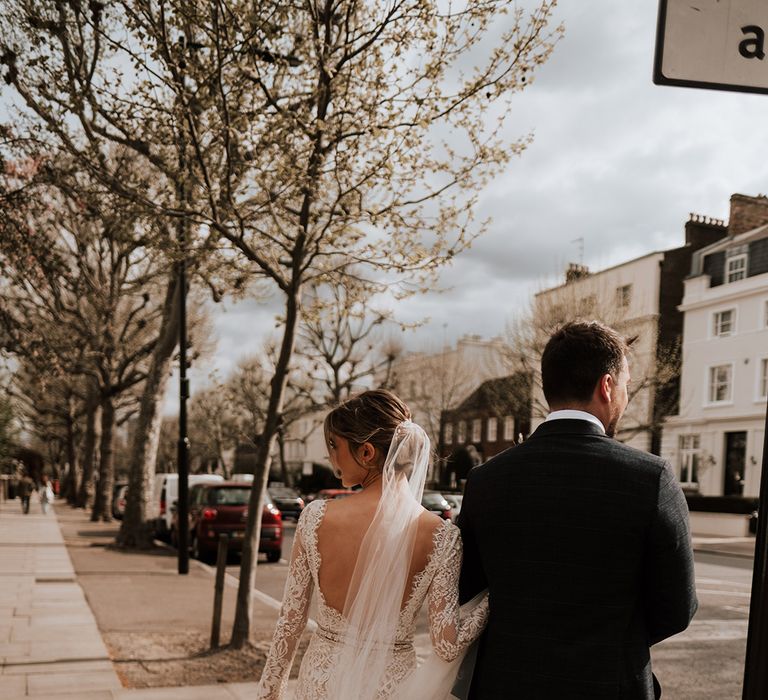 Bride wears low back lace wedding dress with cathedral veil and walks alongside her groom 