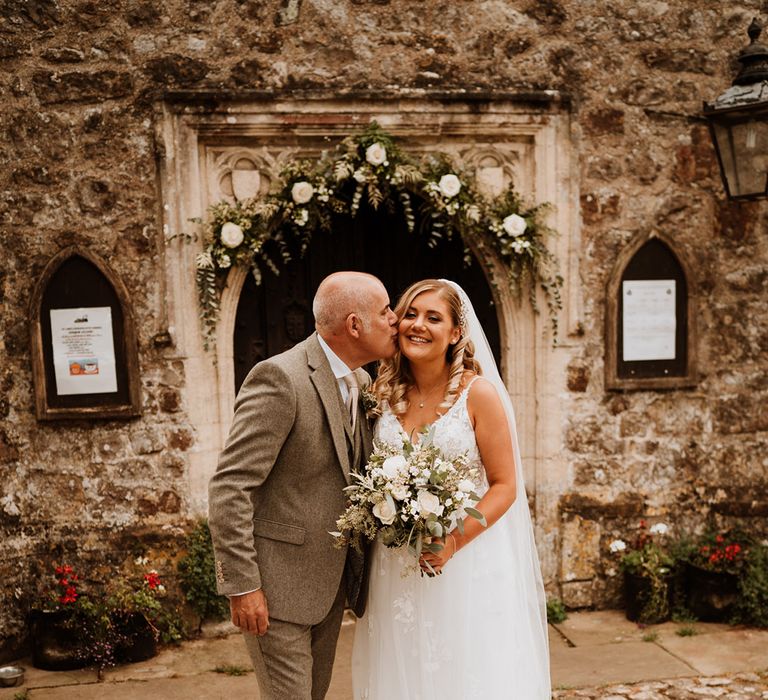 Father of the bride in a dark grey tweed suit kisses the bride on the cheek outside of the church 