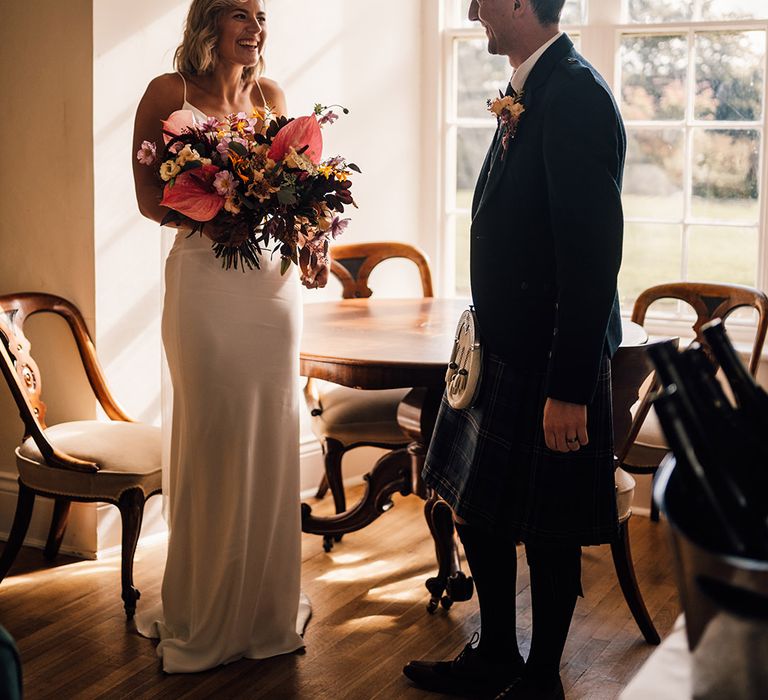 Bride holds tropical bridal bouquet and smiles toward her groom in kilt 