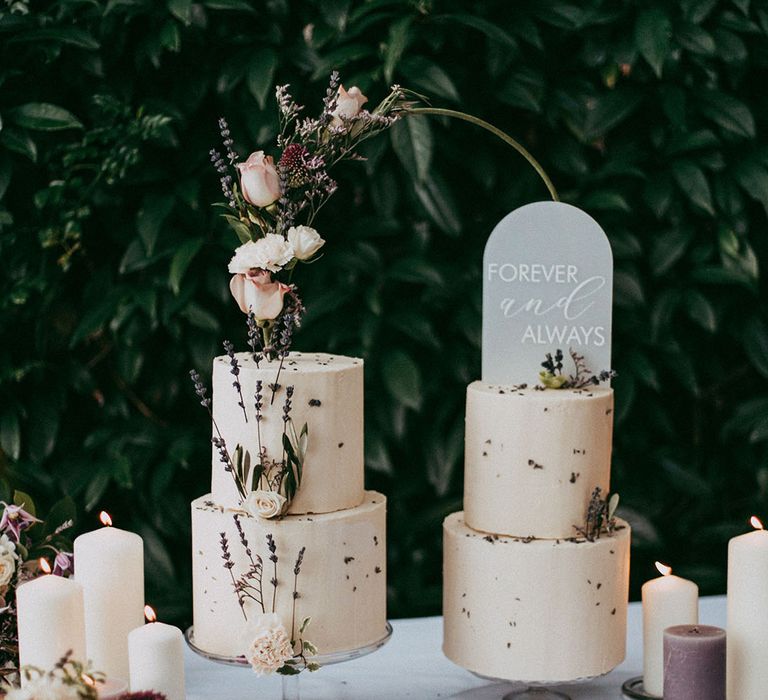 Lavender and white pillar candles standing by two tiered buttercream wedding cakes with white rose and dried lavender decorations and transparent engraved cake topper and dark green half hoop