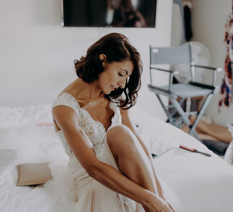 Bride in lace and tulle dress adjust the strap on her gold glittery shoes whilst sitting on bed