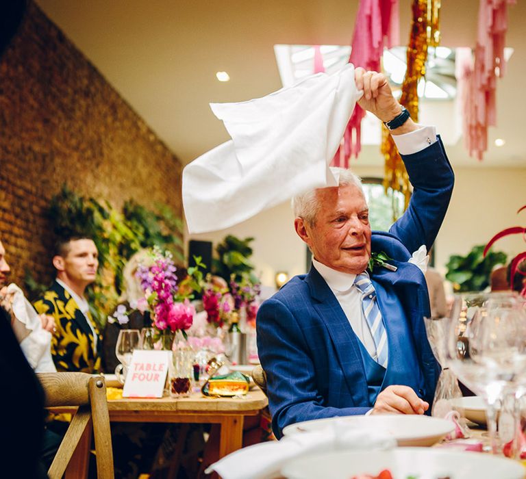 Wedding guest in three piece blue tartan suit with foliage boutonniere waving a napkin in celebration at Middleton Lodge