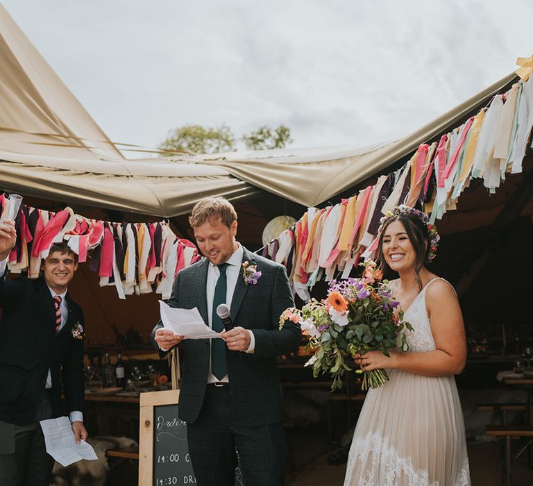 Groom gives speech beside his bride in front of colourful bunting 