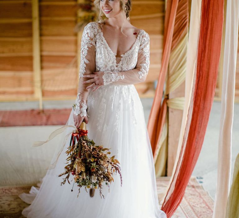 Bride in long sleeved v neck soft ivory dress with botanical embroidered details and headpiece with delicate details and mix of soft gold & bronze metals holding dried autumnal wedding bouquet with white dried cotton stems, terra cotta celosia seeds, green foliage 