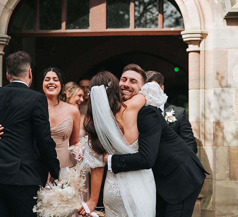 Bride in fitted boho lace wedding dress hugs a groomsman in a black tuxedo 