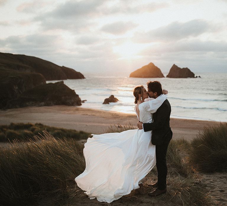 Bride in floaty wedding dress kisses her groom beside the sea during golden hour portraits 