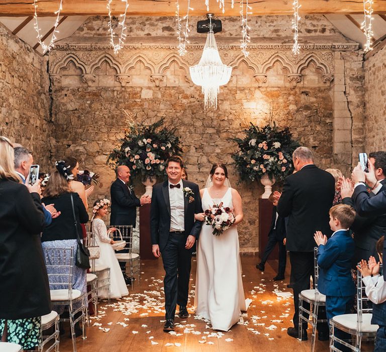 Groom in black tie walk back down the aisle with the bride in a white wedding dress holding a red and white wedding bouquet 