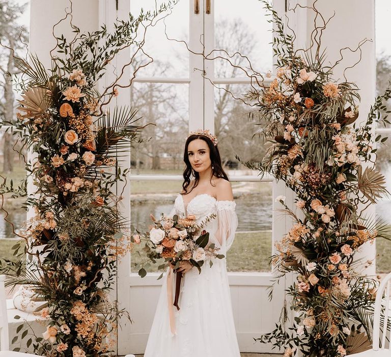 Autumnal wedding flower columns with orange tone flowers and fern leaves with bide holding a matching bouquet and crown in off the shoulder dress