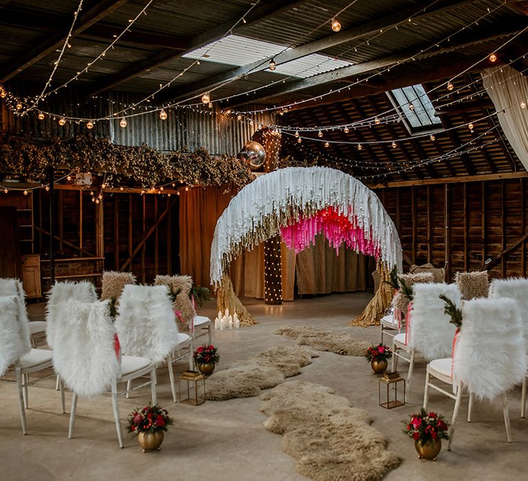 Rustic, boho wedding decor at barn venue - faux fur throws, candles inside lanterns, brass vases with colourful floral arrangements, low crates, cane seating, disco ball wedding decor and large semi-circle arch decor with white, gold and hot pink streamers 