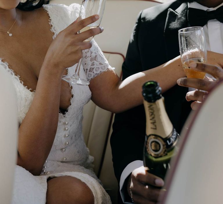 Groom in classic black tuxedo, black bowtie and white pocket square drinking wedding champagne with bride in v neck lace wedding dress 