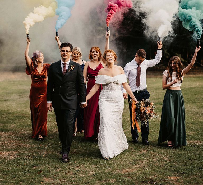 Bride & groom walk alongside wedding party holding colourful smoke-bombs 