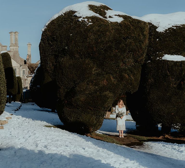 Bride in ruffle bottom wedding dress with white blazer and white heeled shoes holding white flower and foliage bouquet waiting in the snow for the first look moment 