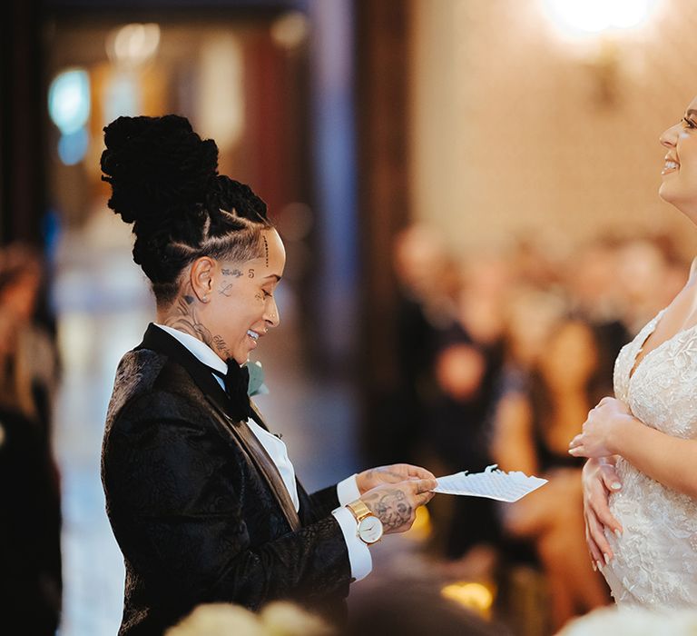 Bride in black snakeskin print tuxedo with black bowtie and white rose boutonniere reading their vows to bride in lace sleeveless v neck Enzoani wedding dress with tulle finish and church length veil holding her baby bump