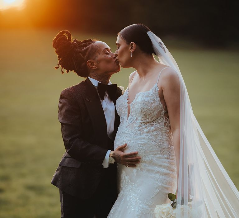 Bride in lace sleeveless v neck Enzoani wedding dress with tulle finish and church length veil and bride in black snakeskin print tuxedo with black bowtie and white rose boutonniere holding her baby bump