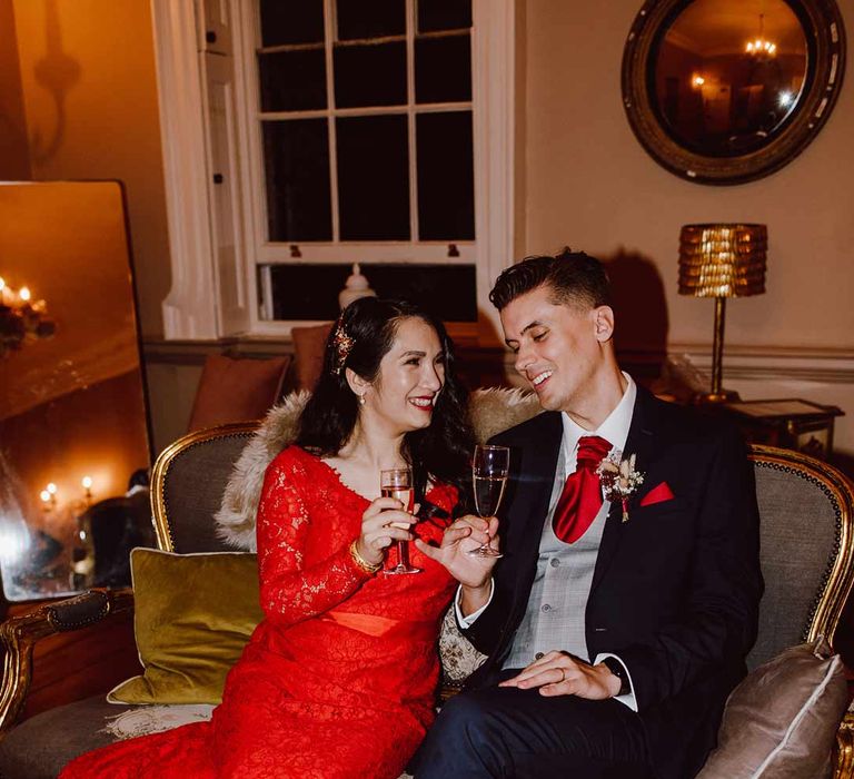 Groom in deep blue grooms blazer, grey waistcoat, red tie, red pocket square and mixed dried flower boutonniere sitting with bride in red lace wedding dress with long sleeves, red and gold hair accessories and golden closed toe wedding heels