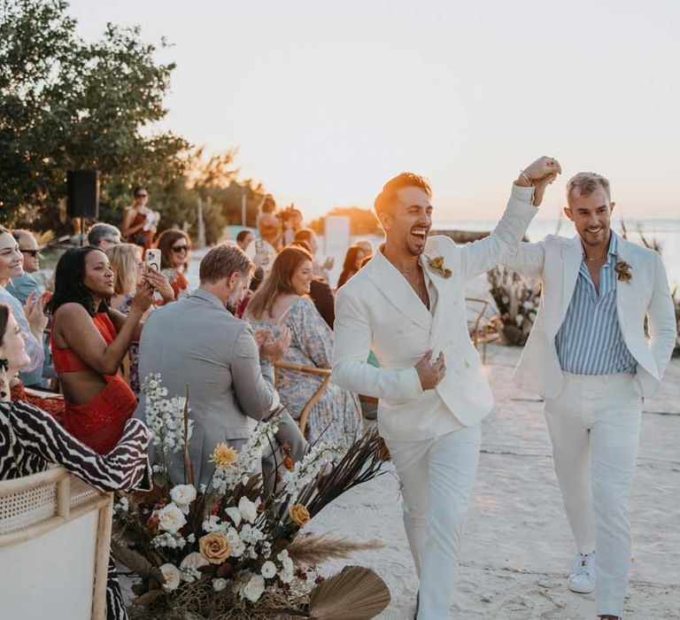 Groom in mens white wedding suit with dusky orange garden rose and dried flower boutonniere, white shirt and silver jewellery holding hands and walking down the aisle with groom in white blazer, light blue and white striped shirt, dusky orange garden rose and dried flower boutonniere and gold jewellery