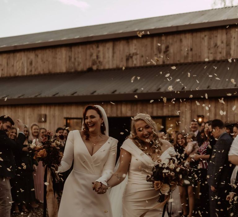 Bride in long sleeve deep v wedding dress and church length veil and bride in satin short sleeve deep v wedding dress and church length veil walking out of Hidden River Cabins to dried rose petal wedding confetti 