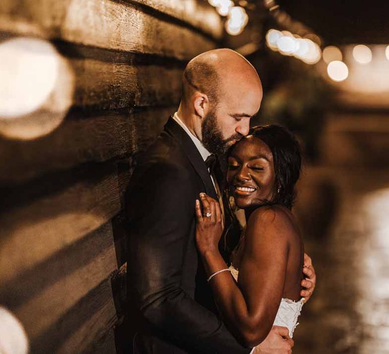 Bride in sweetheart neckline lace wedding dress hugging groom in classic black three piece grooms suit with bow tie and white rose and foliage boutonniere in church at Lains Barn