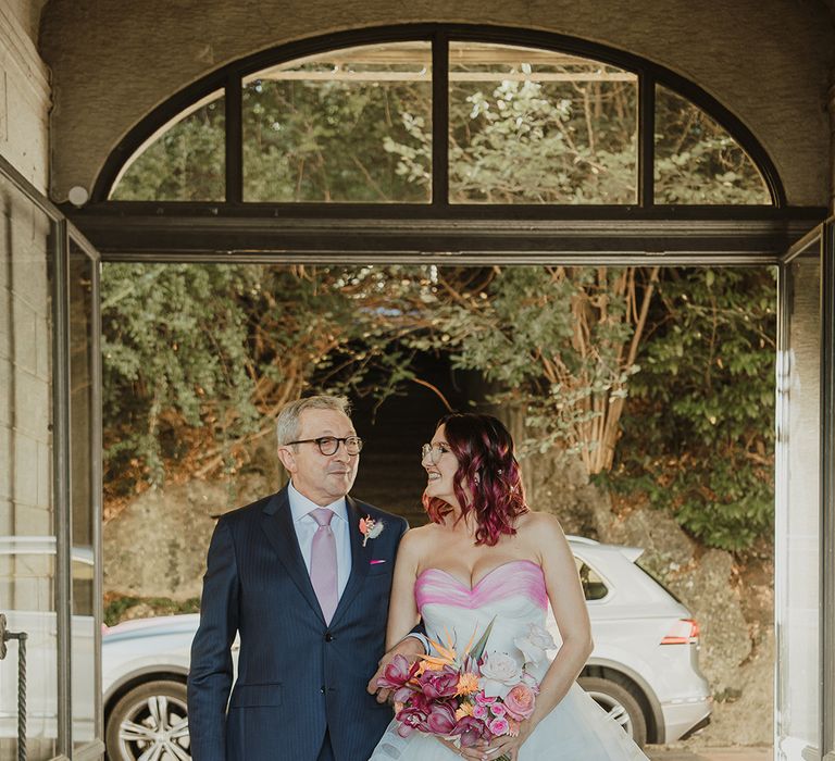 father of the bride in a navy suit and daughter in a strapless wedding dress with layered skirt holding a tropical pink wedding bouquet