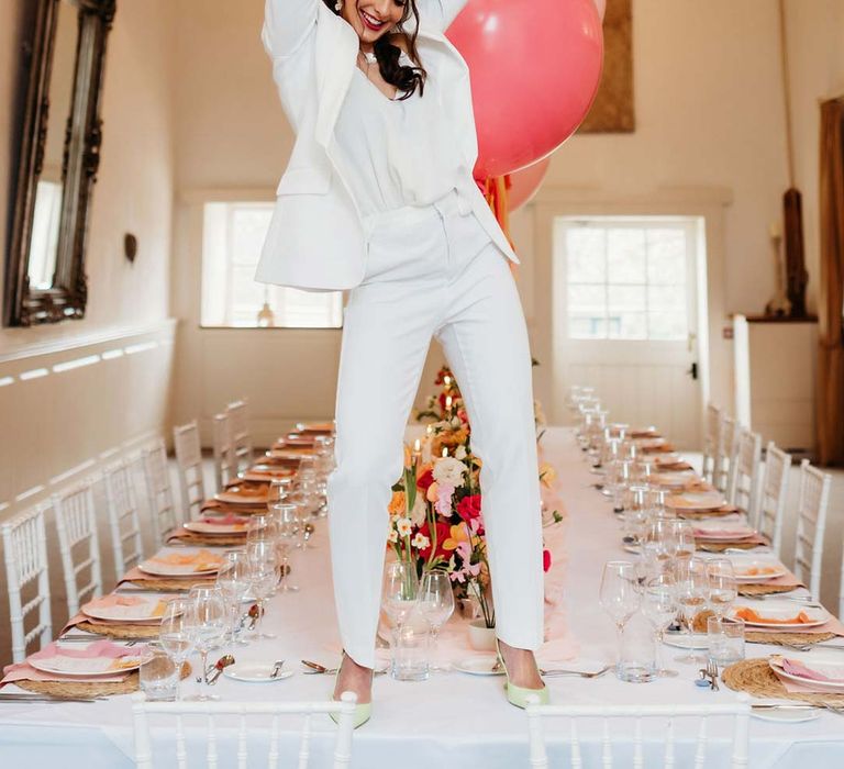 Bride in white bridal suit with closed toe bright green wedding heels standing on wedding tablescape with white wedding tablecloth, burlap place mats, tapered candles, pink and orange wedding stationery, and floral wedding table centrepieces with pink and orange garden roses, orange floribunda, yellow ranunculus flowers, foliage and Iceland poppies 