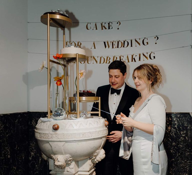 Bride and groom standing by three tiered golden wedding cake stands with three single tier wedding cakes with disco ball toppers and decor, single poppy flowers in test tube vases and golden funny wedding sign