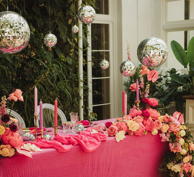 Hot pink wedding theme decor with sweetheart table decorated in pink tablecloth, flowers and candles with hanging disco balls