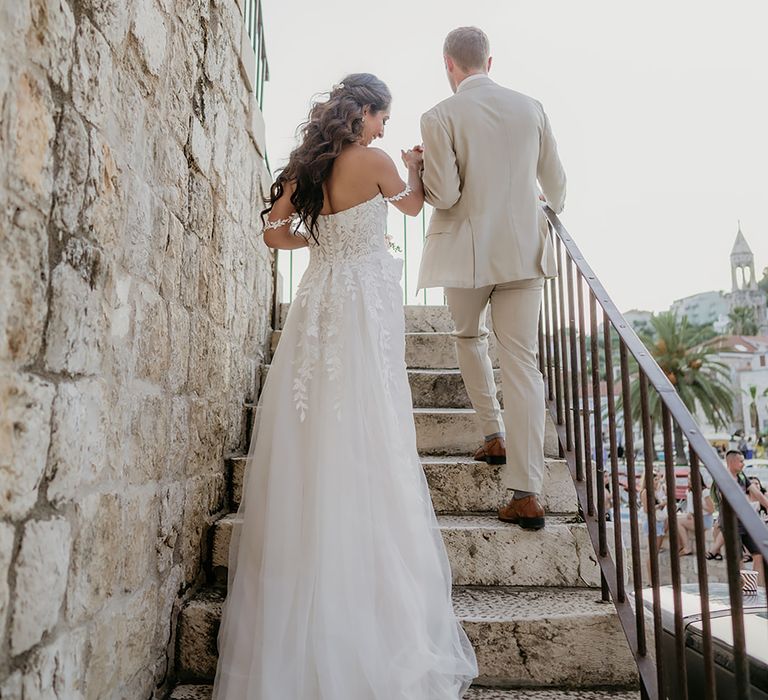 bride in off-the-shoulder-wedding-dress and groom in linen suit walking up stairs in Old Town Hvar