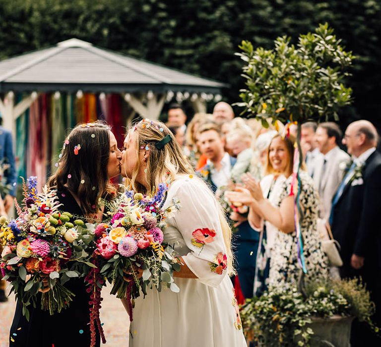 Two brides with one wearing a floral embroidered wedding dress and the other in black bridal jumpsuit share a kiss after their confetti exit 