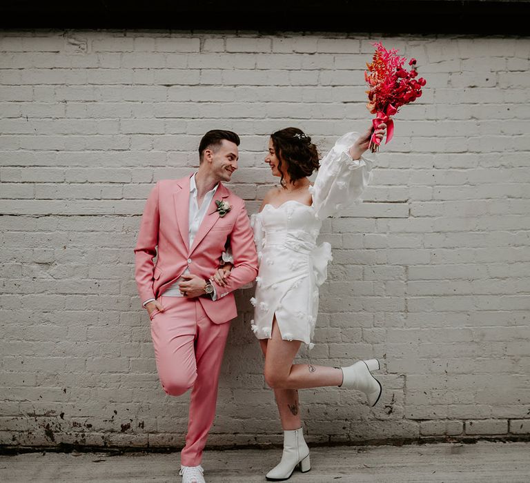 Bride wearing short wedding reception dress with white boots and hot pink wedding bouquet posing with the groom in a pink wedding suit