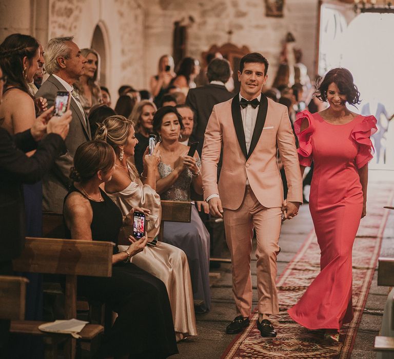 Groom in pink tuxedo walking down the aisle with his mum 