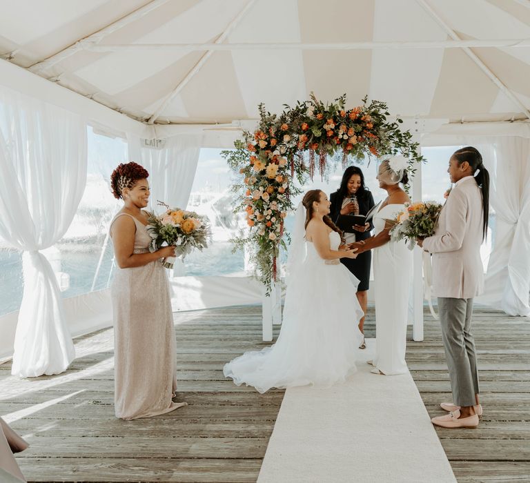 Two brides standing at the orange floral arch altar exchanging vows at their same sex wedding with their best persons by their side 